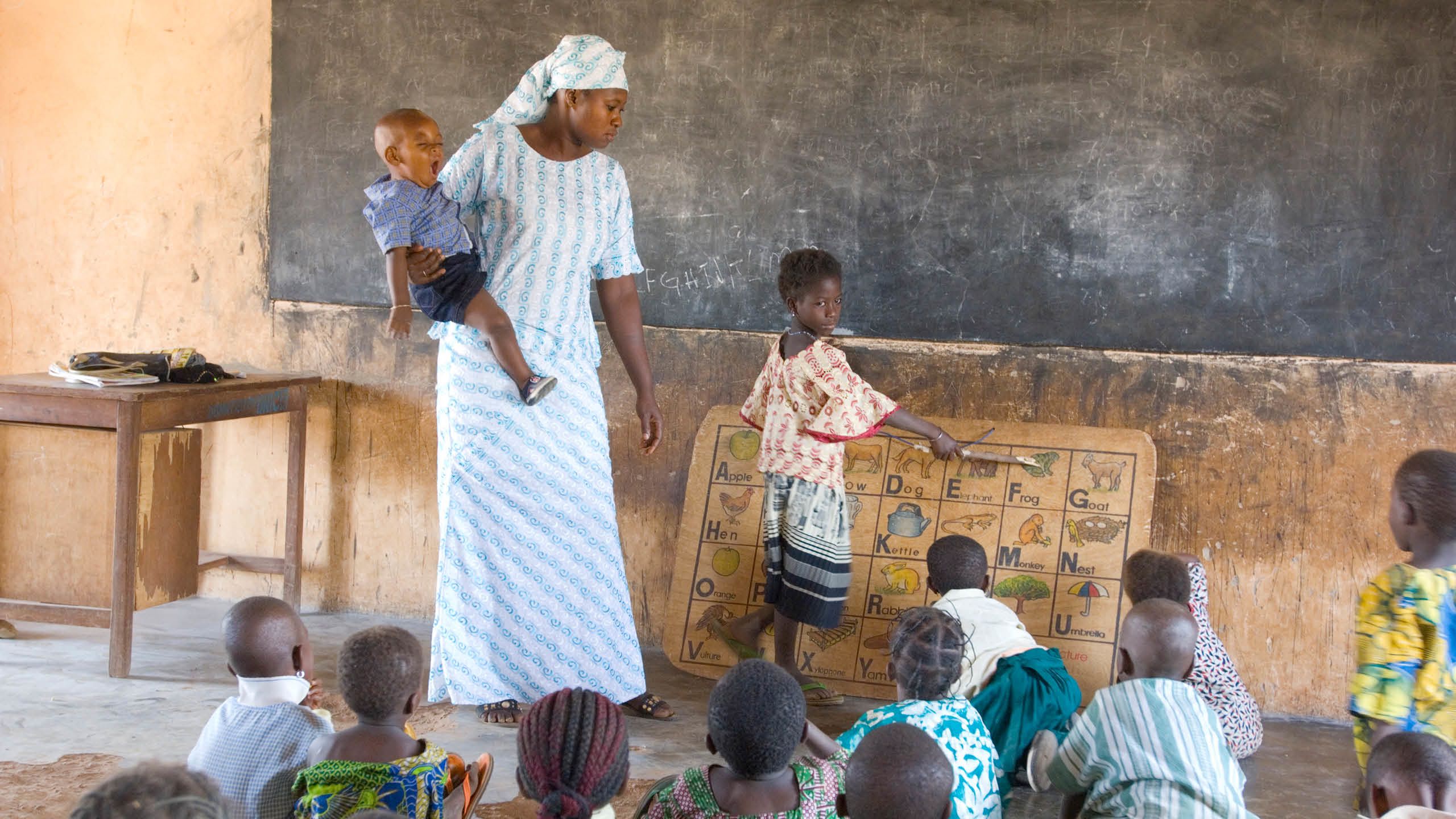 A teacher wearing a matching head scarf and long dress balances a child on her hip while watching a young student point to letters on a wooden board with letters from the alphabet.
